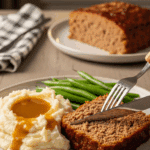 Plated meatloaf slice served with mashed potatoes and green beans at a dinner table.