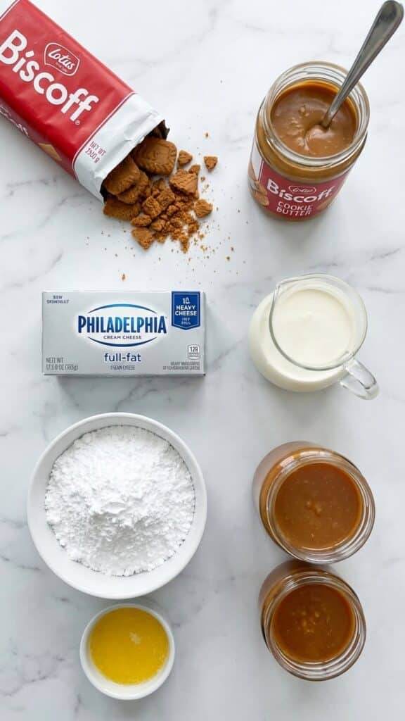 Overhead flat lay view of ingredients for Biscoff cheesecake including Lotus biscuits, jar of Biscoff spread, cream cheese, and heavy cream on a marble table.