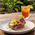 A plate of blackened tuna and mango salsa on an outdoor patio table, with a fork lifting a bite of tuna and salsa, set against a tropical background.