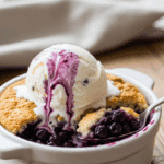 A bowl of warm blueberry cobbler topped with a melting scoop of vanilla ice cream, served in a cozy setting with a spoon lifting a bite.