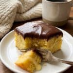 fork taking a bite from a slice of Boston Cream Poke Cake, revealing the pudding-soaked yellow cake and chocolate topping.