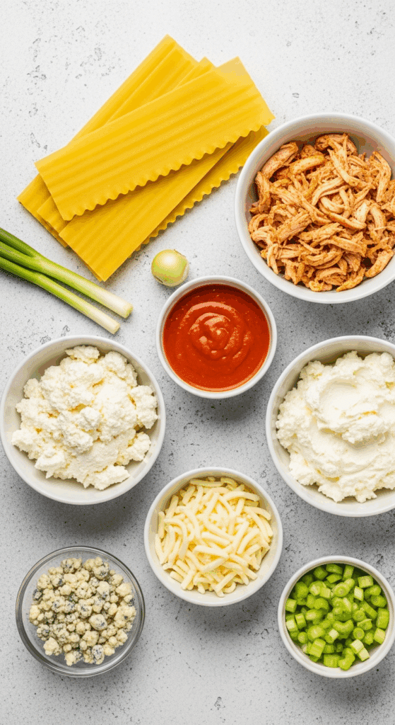 Overhead view of ingredients for buffalo chicken lasagna: lasagna noodles, shredded chicken, buffalo sauce, ricotta mixture, mozzarella, bleu cheese, celery, and green onions arranged on a counter.