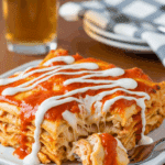 A plated serving of buffalo chicken lasagna drizzled with ranch and hot sauce, with a fork taking a bite, set in a casual dining atmosphere.