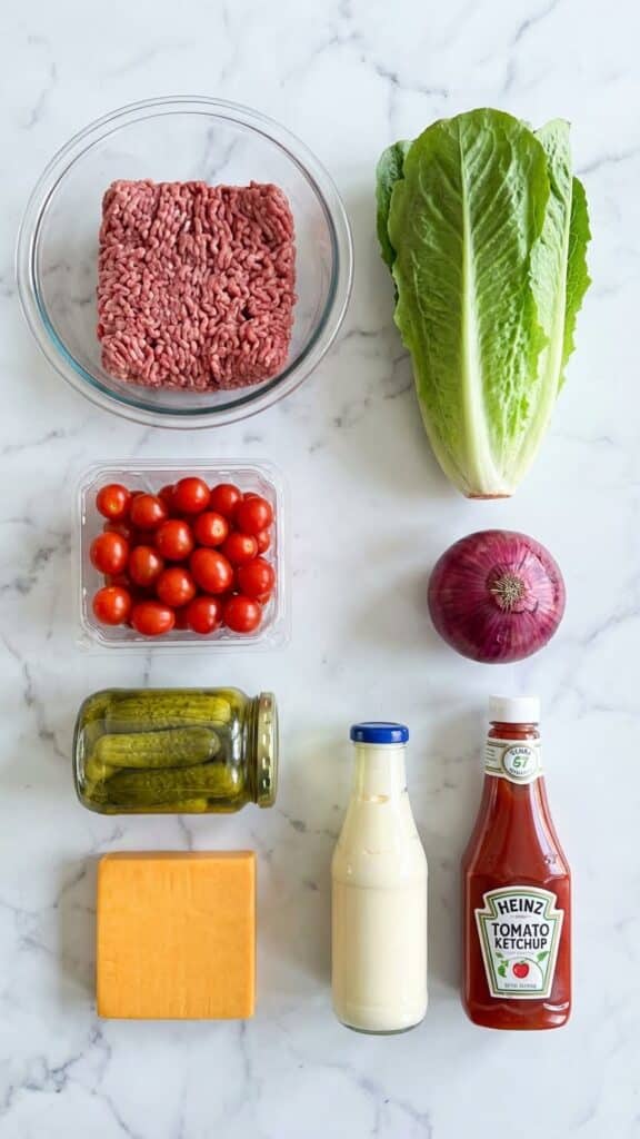 Flat lay of burger bowl ingredients including raw beef, lettuce, tomatoes, cheese, and condiments arranged neatly on marble.