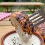 Close-up of a fork dipping a piece of grilled Cajun chicken into a bowl of white sauce.