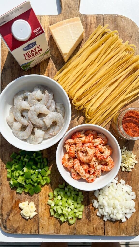 Raw ingredients for Cajun pasta including shrimp, crawfish, pasta, cream, spices, and chopped vegetables on a wooden board.