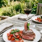 A plate of grilled steak and caprese salad served with a glass of red wine on an outdoor table.