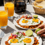 Two plates of carnitas huevos rancheros served on a sunny patio table with orange juice and coffee in the background.