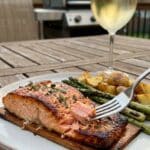 A plated serving of cedar plank salmon with grilled asparagus and potatoes on an outdoor patio table, with a fork lifting a bite.