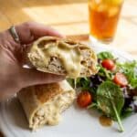 A hand holding a halved cheesy garlic chicken wrap over a plate with a side salad, ready for lunch in a sunlit kitchen.