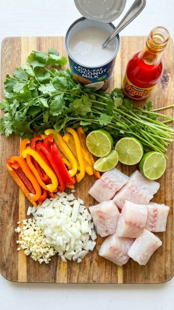 Overhead view of ingredients for Moqueca: raw fish chunks, colorful peppers, lime, cilantro, coconut milk, and a bottle of dendê oil on a wooden board.