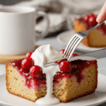 A slice of warm cherry upside-down bread pudding with whipped cream on a plate, with a fork lifting a bite in a cozy setting.