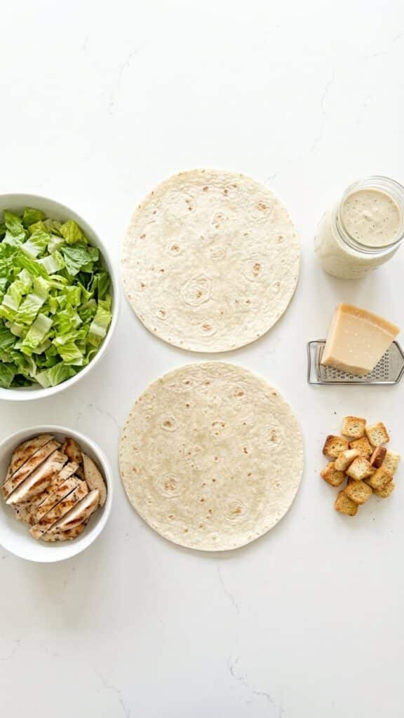 Flat lay of ingredients for chicken Caesar wraps including tortillas, lettuce, chicken, and dressing.
