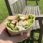 A hand holding a chicken Caesar wrap with a bite taken out of it.