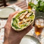 A close-up POV shot of a hand holding a fresh Chicken Caesar Wrap half, about to take a bite.