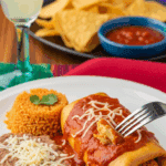 A plate with a large chimichanga covered in sauce, served with Mexican rice and beans, with a fork cutting into it in a festive dining setting.