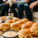 A platter of warm Chicken Cordon Bleu sliders served with a side of Dijon sauce at a party, with people mingling in the background.