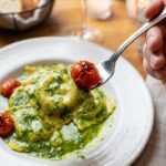 A close-up POV shot of a fork lifting a pesto-covered chicken ravioli and a burst cherry tomato.
