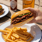 A hand holding a chipotle beef sandwich on a plate with french fries, set on a picnic table with a beer in the background.