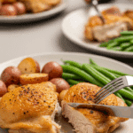 Plated crispy baked chicken thighs served with roasted potatoes and green beans on a dinner table, with one thigh being cut to show juiciness.