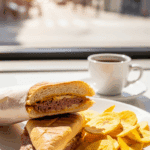 A pressed Cuban sandwich served with plantain chips on a plate, with a cup of Cuban coffee in the background at a sunlit cafe table.