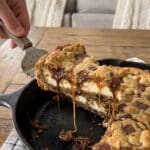 A massive slice of deep dish cookie dough cheesecake pizza being lifted by a server, revealing a thick cheesecake layer sandwiched between cookie dough layers.