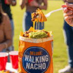 A hand holding a loaded walking nacho bag with a fork lifting a bite, set against a background of an outdoor tailgate party.