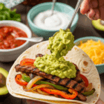 A hand holding a flour tortilla filled with fajita filling, being topped with guacamole, set against a background of various toppings on a dining table.