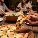 A hand holding a folded slice of cheesy Margherita pizza at a dinner table, with the rest of the pizza and glasses of wine in the background.
