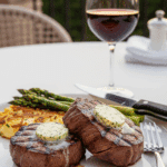 Plated slices of grilled beef tenderloin with melting herb butter, asparagus, and potatoes on an outdoor dining table.