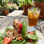 A fork holding a bite of chicken and strawberry salad with an outdoor background.