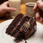 A close-up of a fork taking a bite from a slice of moist Hershey's chocolate cake with fudge frosting on a plate.