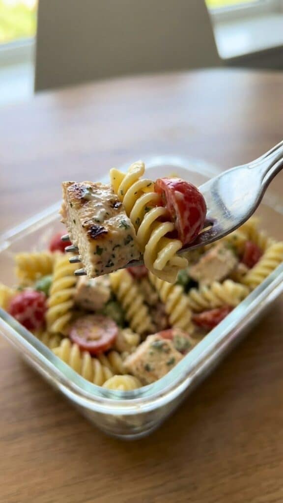 Close-up of a forkful of creamy chicken and pasta salad with a meal prep container in the background.