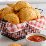 A basket of baked pizza pockets served with a side of marinara dipping sauce on a kitchen counter.