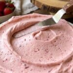 Close-up of a spatula spreading fluffy pink strawberry buttercream onto a cake.