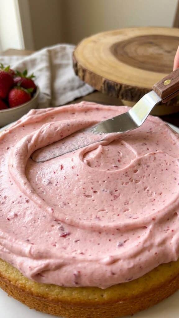 Close-up of a spatula spreading fluffy pink strawberry buttercream onto a cake.