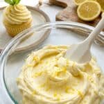 Close-up of an offset spatula spreading thick, creamy lemon frosting onto a yellow cake layer, showing lemon zest specks.