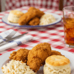 Plated fried chicken served with coleslaw and a biscuit on a checkered tablecloth setting.