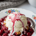 A bowl of warm cherry cobbler topped with a melting scoop of vanilla ice cream, served in a cozy setting with a coffee mug in the background.