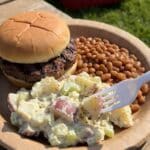 A scoop of potato salad served on a plate with a burger and baked beans at a BBQ.