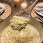 A close-up POV shot of a serving spoon scooping up hot, creamy lemon orzo pasta with steam rising.