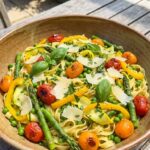 A close-up POV shot of a fork twirling tagiatelle pasta mixed with asparagus, tomatoes, and peas.