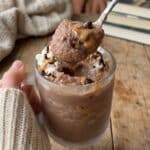 A close-up of a spoon scooping up thick, icy peanut butter chocolate slush from a glass held by a hand in a sweater.