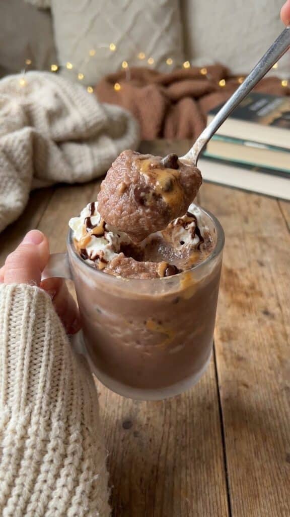 A close-up of a spoon scooping up thick, icy peanut butter chocolate slush from a glass held by a hand in a sweater.