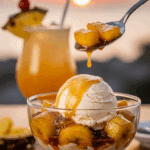 A glass bowl of warm bananas and pineapple foster with ice cream, with a spoon lifting a bite, set against a sunset cocktail background.