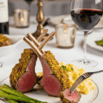 Plated pistachio lamb chops with potato gratin and asparagus on an elegant dinner table with wine.