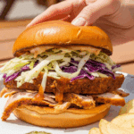 A hand holding a pulled turkey tenderloin sandwich on a brioche bun topped with coleslaw, set in a casual outdoor dining scene.