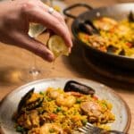 A plate of quick paella with a hand squeezing a lemon wedge over it, set in a casual dinner atmosphere with wine in the background.