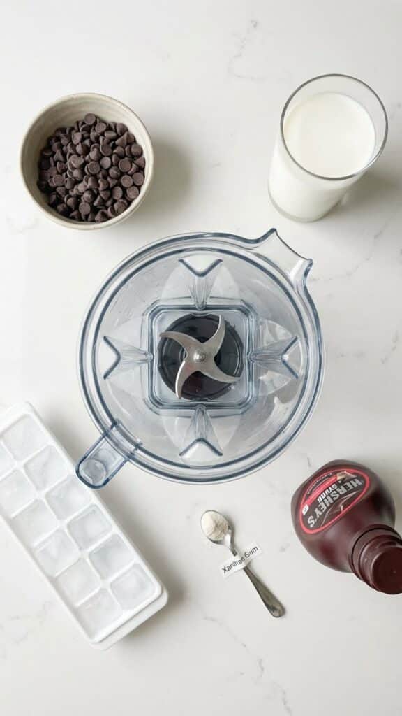 Ingredients for a chocolate frappuccino including milk, ice, chocolate chips, and blender.