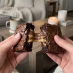 A close-up of hands pulling apart a warm Reese's brownie, showing a gooey, stretchy melt of chocolate and peanut butter in the center.